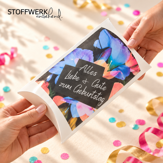 STOFFWERK Geschenkgutschein in Kissenverpackung mit buntem Blumenmotiv und Geburtstagsgruß, von zwei Händen überreicht; nach Originalfotos inszeniert.
