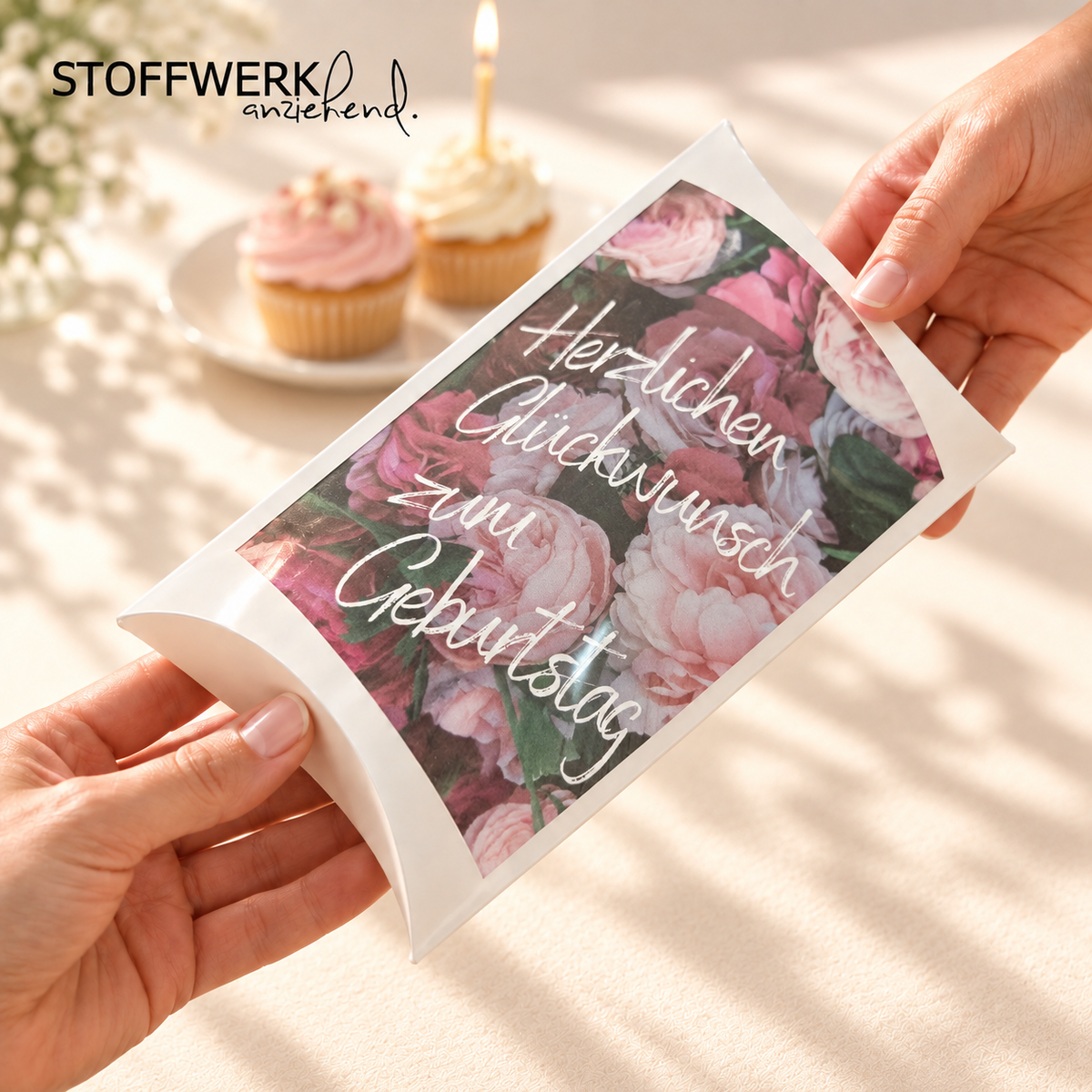 STOFFWERK Geburtstagsgutschein in gewölbter Kissenverpackung mit rosafarbenem Rosenmotiv, von zwei Händen überreicht. Zwei Muffins und eine Kerze im Hintergrund; nach Originalfotos inszeniert.